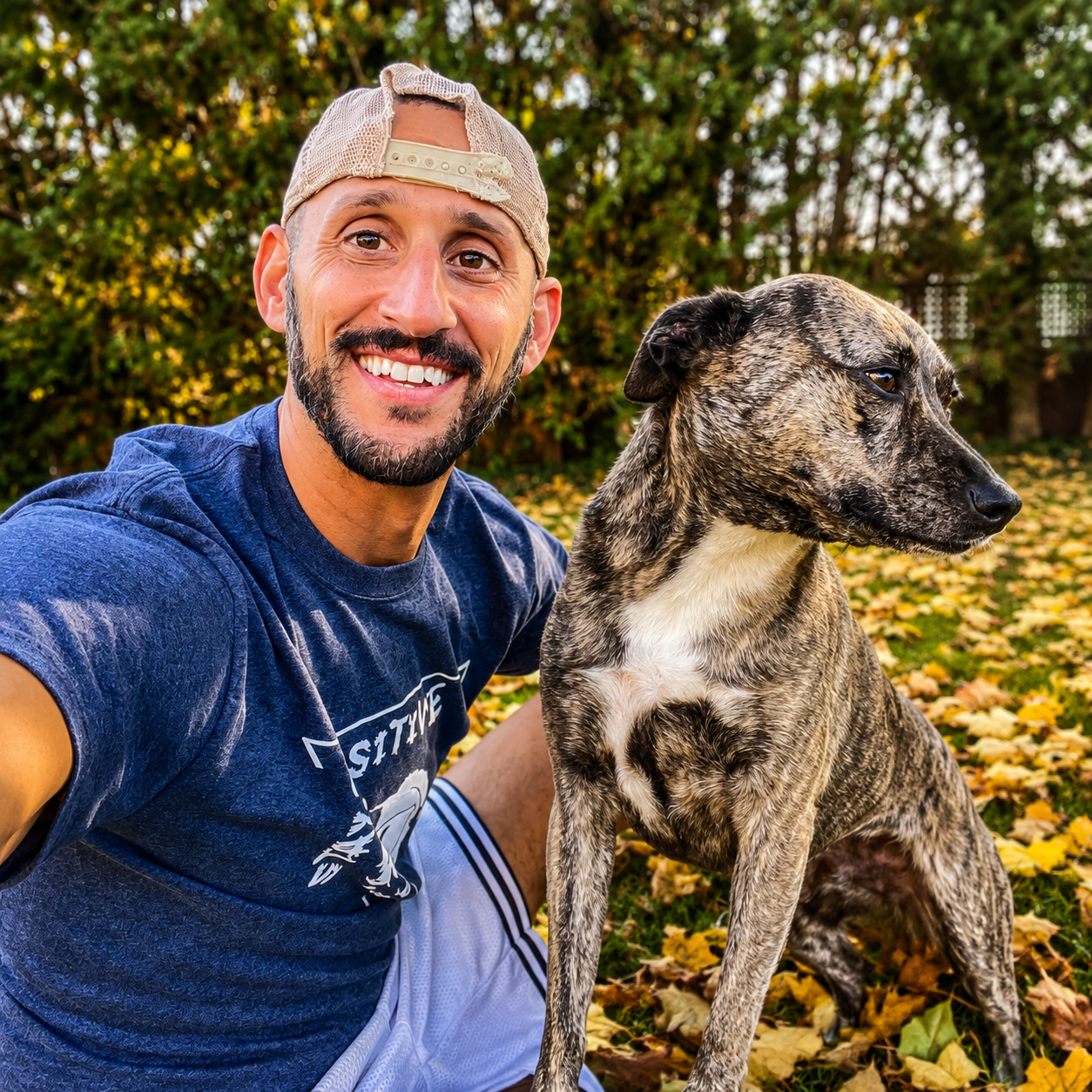 Joseph smiling beside his brindle rescue dog among fallen autumn leaves
